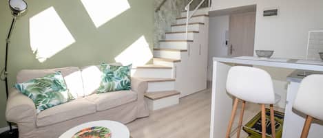 A modern living space with a staircase, a sofa with tropical pillows, a dining area with bar stools, and a kitchen counter.