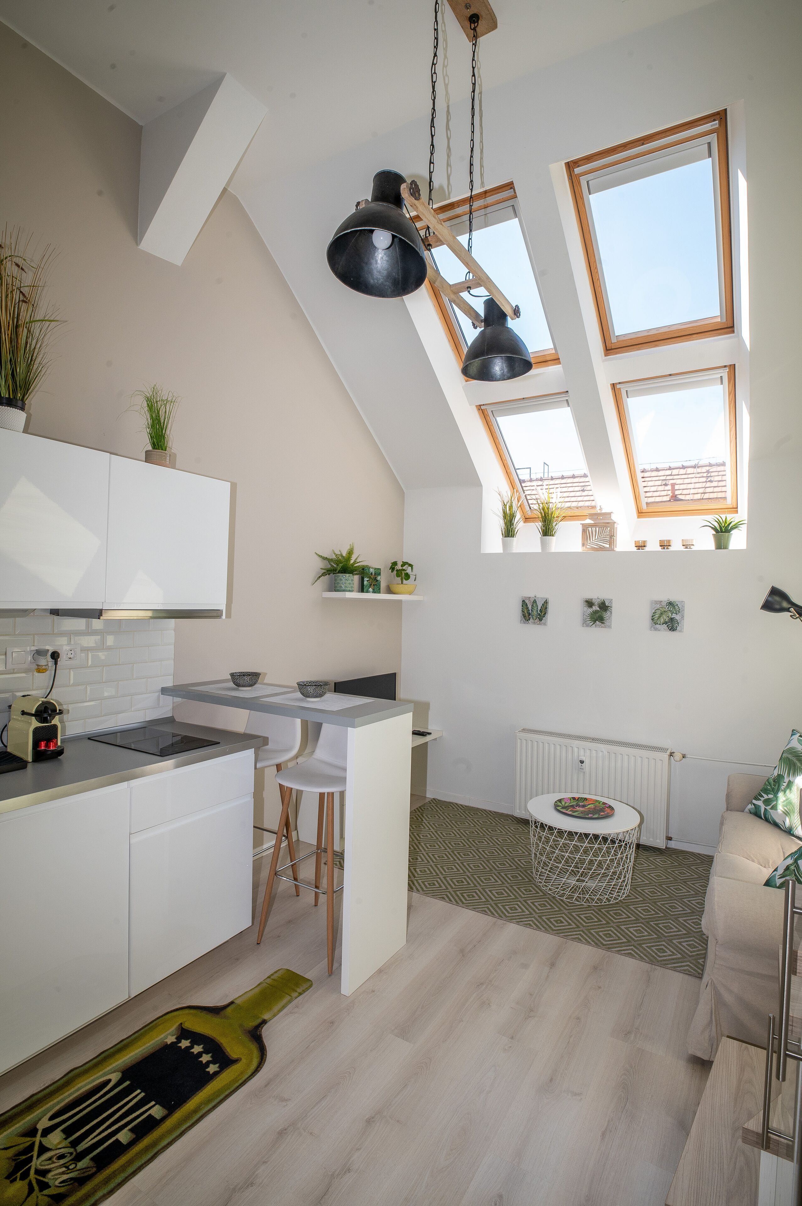 Une cuisine moderne avec des armoires blanches, un comptoir noir et un puits de lumière. On y trouve des plantes sur le rebord de la fenêtre, ainsi qu’une table ronde entourée de chaises.
