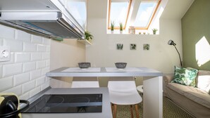 A compact kitchen with a central island, white tiled backsplash, and a built-in stove. There are two white chairs, a sofa with green cushions, and a small table with a lamp. - Palms Apartments Budapest (Budapest)