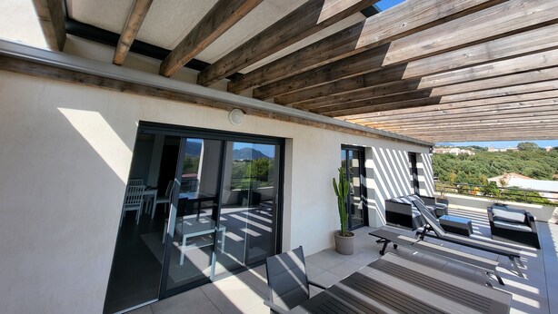 A modern outdoor patio with wooden beams, a glass door leading to a seating area, and a view of the surrounding landscape. - 6/8 person apartment, large terrace (Porto-Vecchio)