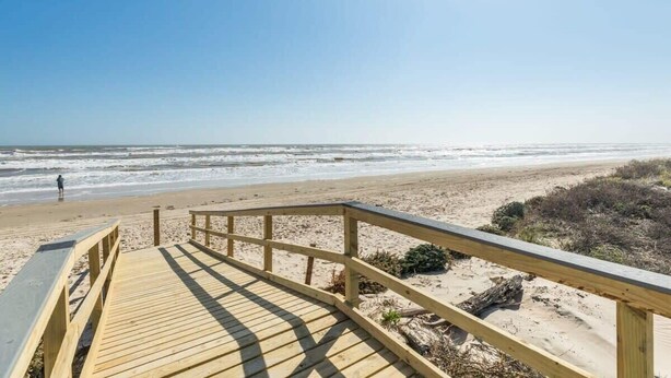 Ein hölzerner Steg führt zu einem Strand bei blauem Himmel.