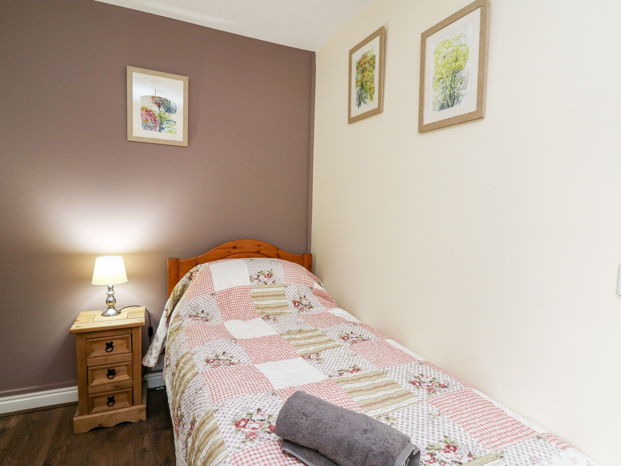 A bedroom with a wooden bed, a bedside table with a lamp, and two framed pictures on the wall.