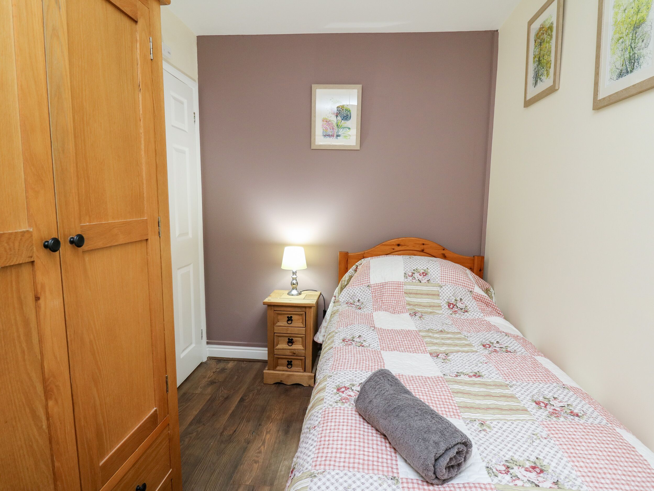 Un dormitorio con un armario de madera, una mesita de noche con lámpara y una cama con una toalla enrollada.