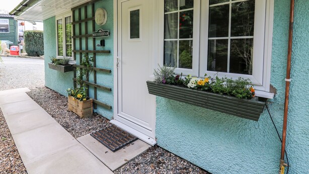 Una cabaña con puerta blanca, ventana de cuadrícula y pequeño porche con banco y plantas en macetas.