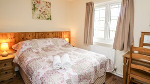 A bedroom with a wooden headboard, a floral bedspread, a bedside lamp, a window with curtains, and a wooden chair. - Primrose Cottage (Llandrindod Wells)
