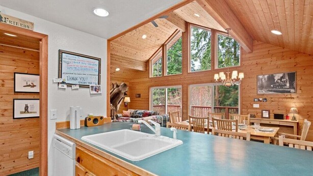 A cabin interior with a kitchen area, wooden walls, a dining table, and a large window. - Cabin Near Leavenworth - Deck + Pets Free (Leavenworth)