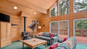 A log cabin interior with a fireplace, plaid furniture, and a large window overlooking a deck. - Cabin Near Leavenworth - Deck + Pets Free (Leavenworth)