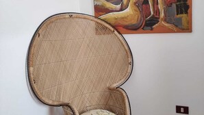 A wicker chair with a cushion, a painting on the wall, and a tiled floor. - Apartment (Santa Teresa Gallura)