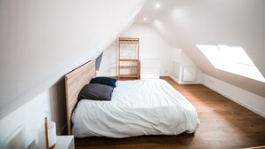 Un dormitorio con techo inclinado, cama con ropa de cama blanca, cabecera de madera, cómoda blanca, armario de madera y tragaluz.