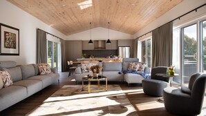 Un salón amplio con un sofá seccional grande, una mesa de centro redonda y un sillón cómodo. Se ve una cocina con salpicadero de ladrillo y electrodomésticos modernos a través de una puerta abierta.