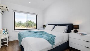 Ein Schlafzimmer mit großem Fenster, einem Bett mit blauer Decke, einem Nachttisch mit Lampe und einer weißen Kommode.
