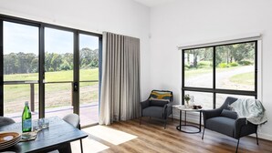 Ein Zimmer mit einer großen Schiebeglasdoor, einem kleinen runden Tisch, zwei Stühlen, einem Sofa und Blick auf eine grüne Landschaft.