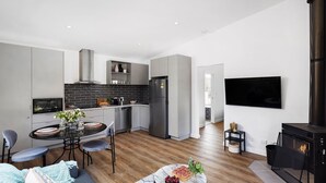Une cuisine moderne avec des meubles gris, un dosseret de carreaux noirs et des appareils électroménagers en acier inoxydable. On y trouve un coin repas avec une table et des chaises, ainsi qu’un salon doté d’une cheminée et d’un téléviseur à écran plat.