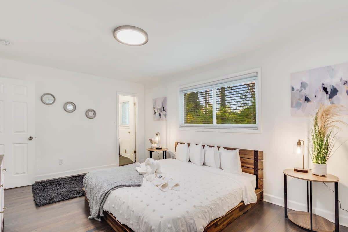 A bedroom with a large bed, a bedside table, a window with blinds, a door, and a wall-mounted light fixture.