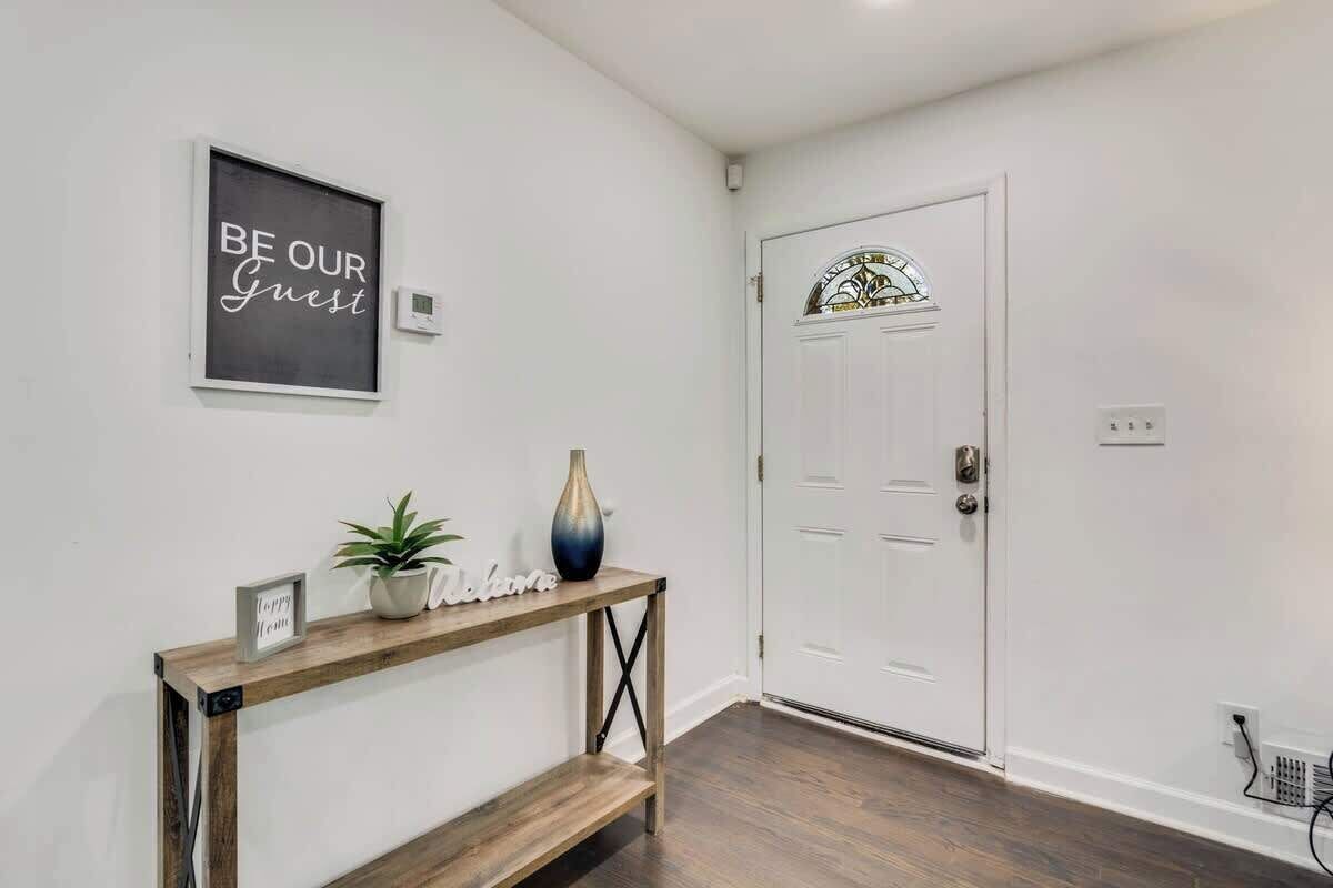 A hallway with a white door, a welcome sign, and a small table with a potted plant and a vase.