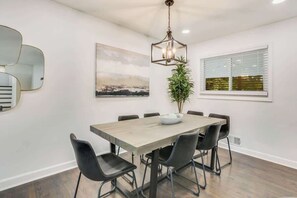A dining area with a rectangular table, six chairs, a hanging light fixture, a potted plant, and a window with blinds.