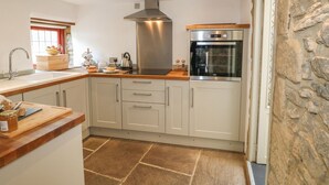 A modern kitchen with white cabinets, a stainless steel range hood, a built-in oven, and a stone wall. - Hope Cottage (Bakewell)