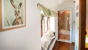 A hallway with a framed painting of a rabbit, a bench with pillows, and a shower area with a glass door. - Hope Cottage (Bakewell)