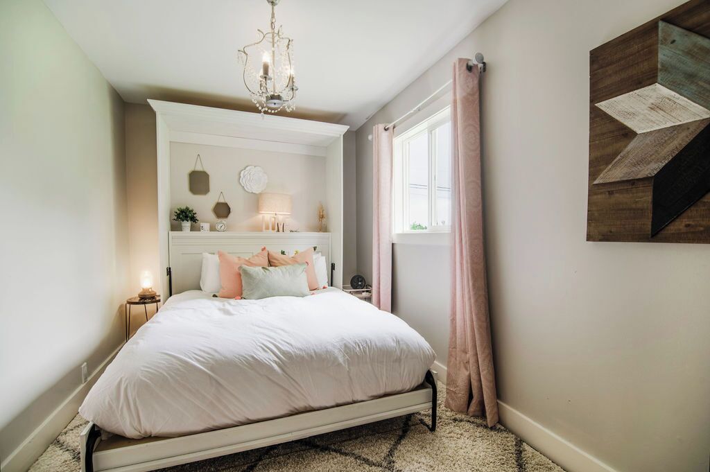 A bedroom with a bed, a chandelier, a window with curtains, and a wall-mounted shelf with decorative items.