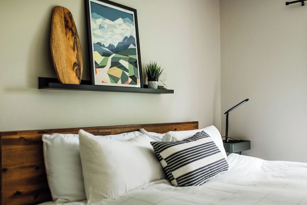 A neatly made bed with white bedding and striped pillows, a wooden headboard, a framed abstract painting, a wooden object on a shelf, and a small potted plant.