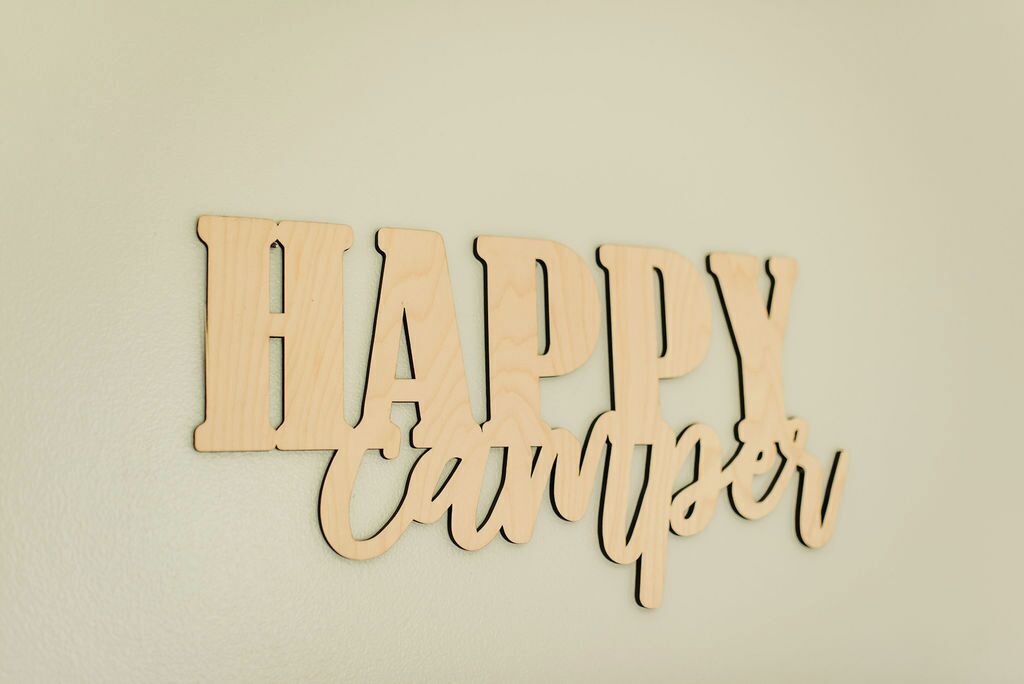 Et træskilt med ordene "HAPPY Camper" på en ensfarvet væg.