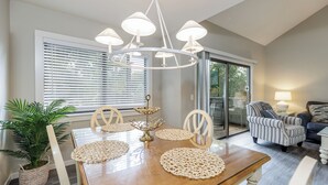 Uma área de refeições com mesa de madeira, cadeiras e um lustre. Há uma planta em vaso e uma janela com persianas.