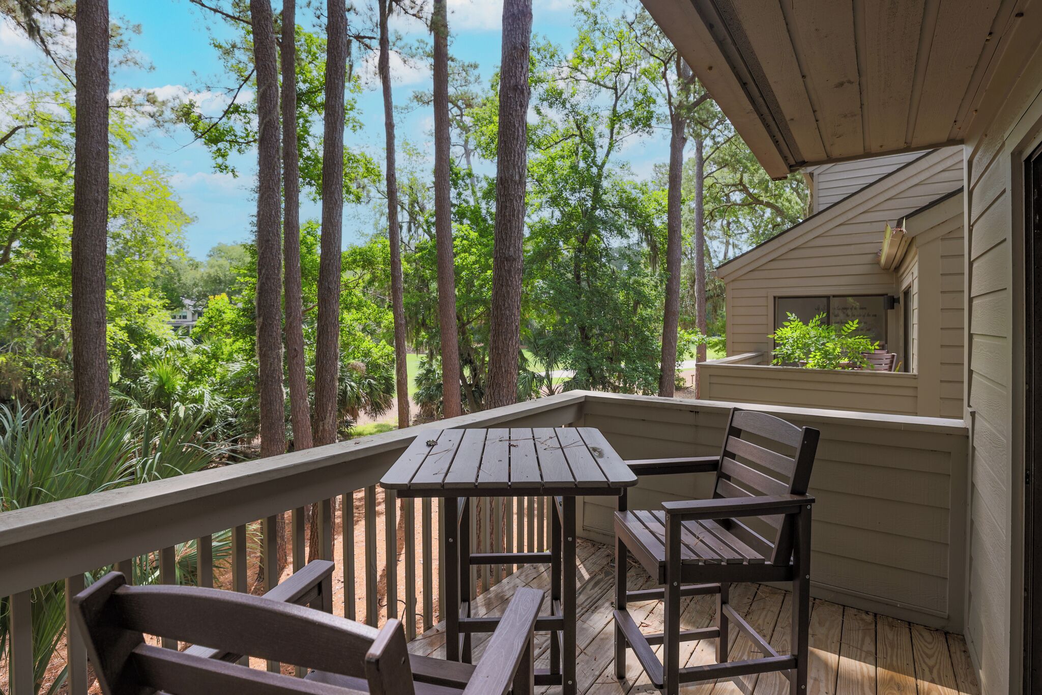 A wooden deck with a table and chairs, surrounded by tall trees and a house in the background.