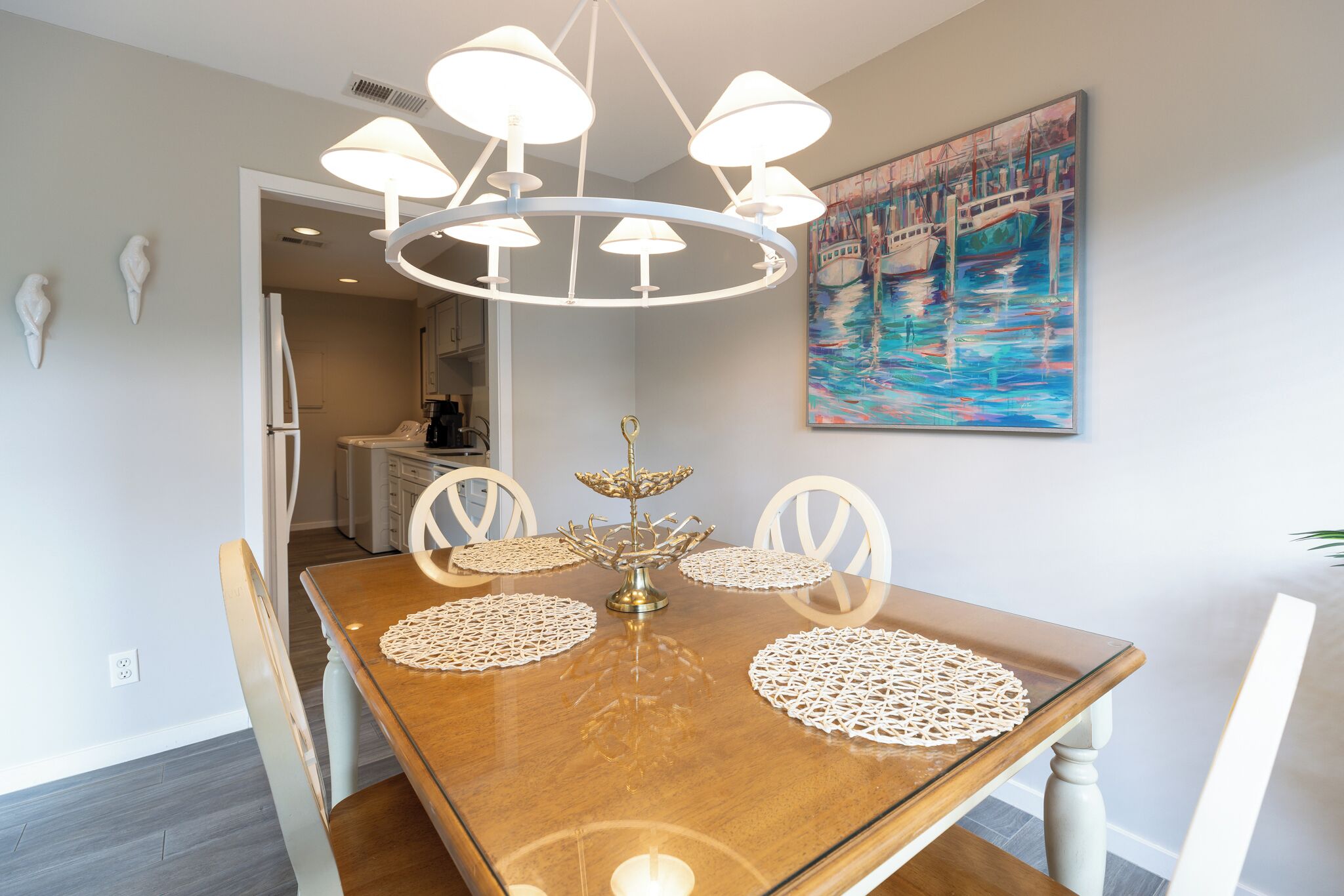 A dining area with a wooden table, white chairs, a chandelier, and a painting of boats on the wall.