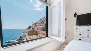 A bedroom with a large window overlooking a coastal town, a bed, a dresser, and a flat-screen TV.