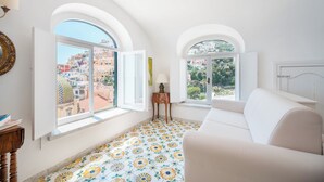 A bright room with a white sofa, a small table, and a lamp. There are two windows with arched tops, and the floor has a floral tile pattern.