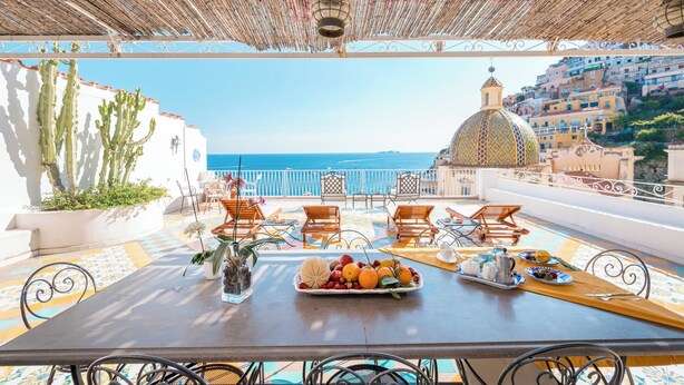 An outdoor dining area with a table set for two, overlooking a coastal view with buildings and the sea.