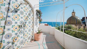A balcony with a tiled wall featuring a floral pattern, a potted plant, and a view of the sea and a dome-shaped building.