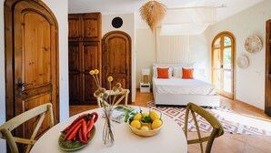 Quarto com cama, mesa de cabeceira e janela. Possui também um espaço de refeições com mesa e cadeiras.
