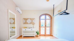 Quarto com cama, penteadeira branca, espelho, ar condicionado fixado na parede, porta de madeira e luminária suspensa.