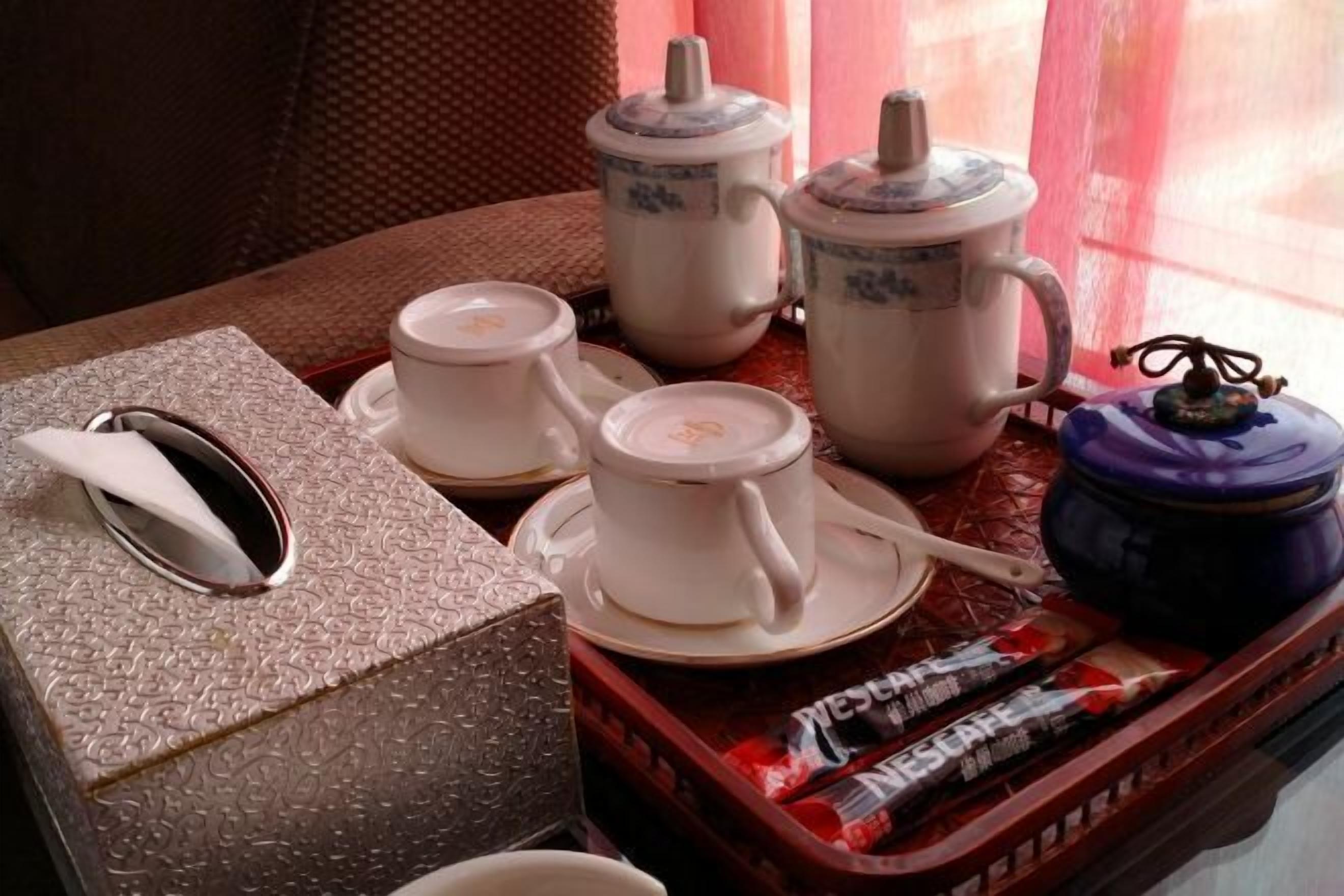 Una mesa dispuesta con tazas de té blancas, platillos, una tetera, una caja de pañuelos y una bandeja con bocadillos empaquetados.