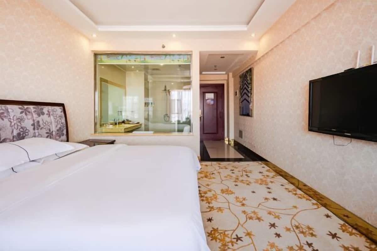 Ein Hotelzimmer mit einem großen Bett, einem Fernseher und einem durch eine Glastür einsehbarer Badezimmer.