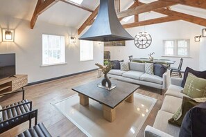 Ein großzügiges Wohnzimmer mit hohen Decken, Holzbalken, einem zentralen Couchtisch, einer Couch und einem Fernseher.