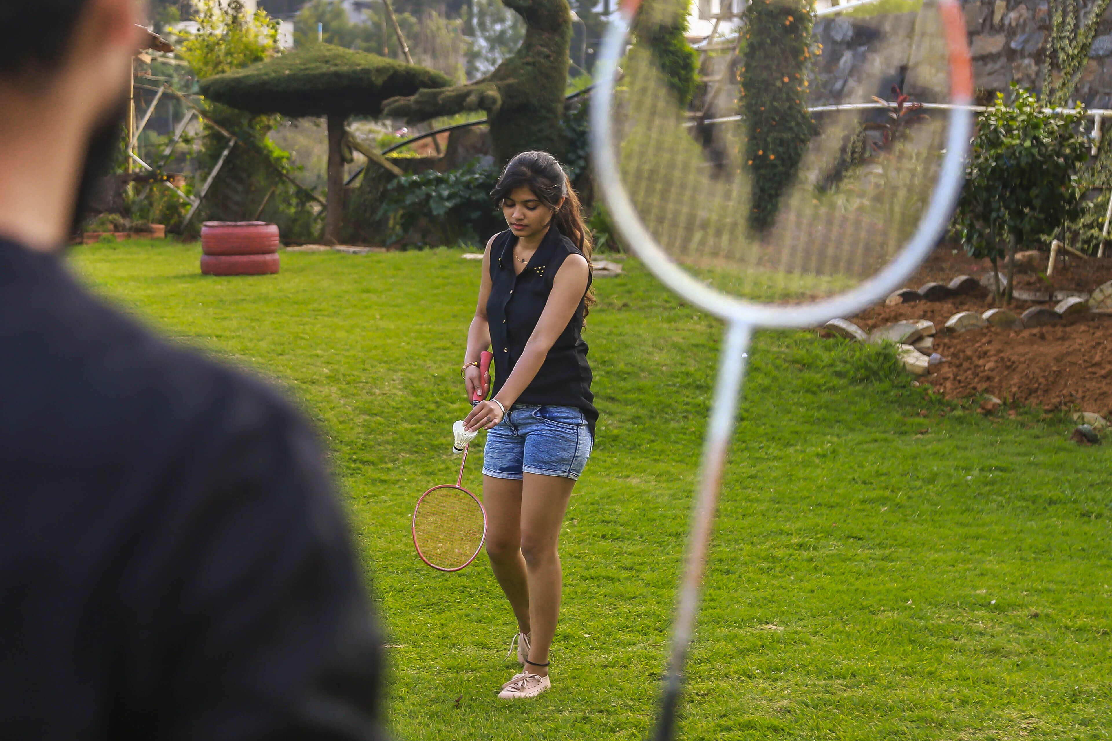 Una persona sosteniendo una raqueta de tenis en un área exterior con césped, una pared de piedra y árboles al fondo.
