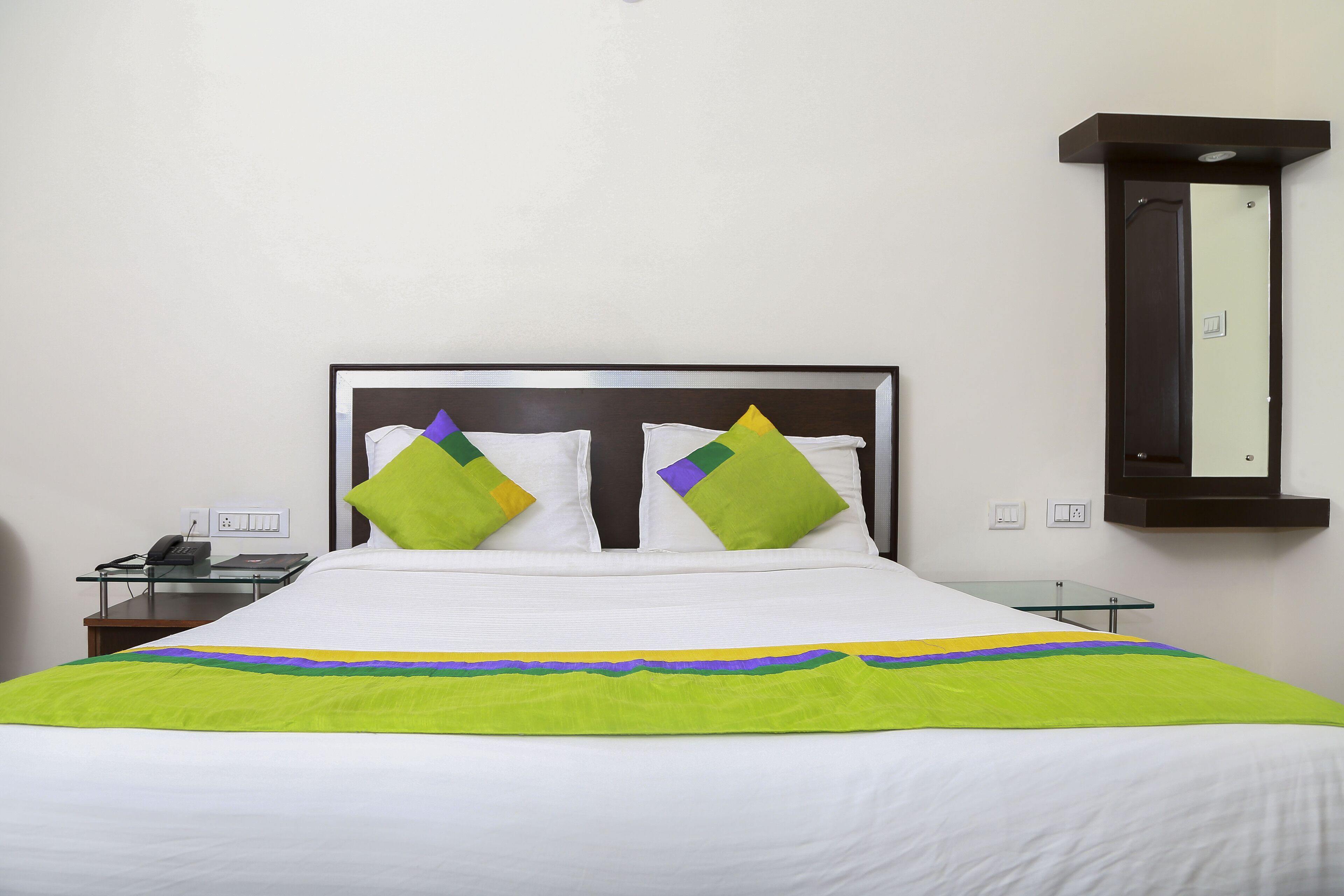 Cama bem arrumada com roupa de cama branca e almofadas verdes e roxas, mesa de apoio de vidro com um telefone e espelho fixado na parede.