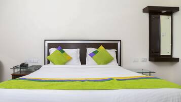 Cama bem arrumada com roupa de cama branca e almofadas verdes e roxas, mesa de apoio de vidro com um telefone e espelho fixado na parede.