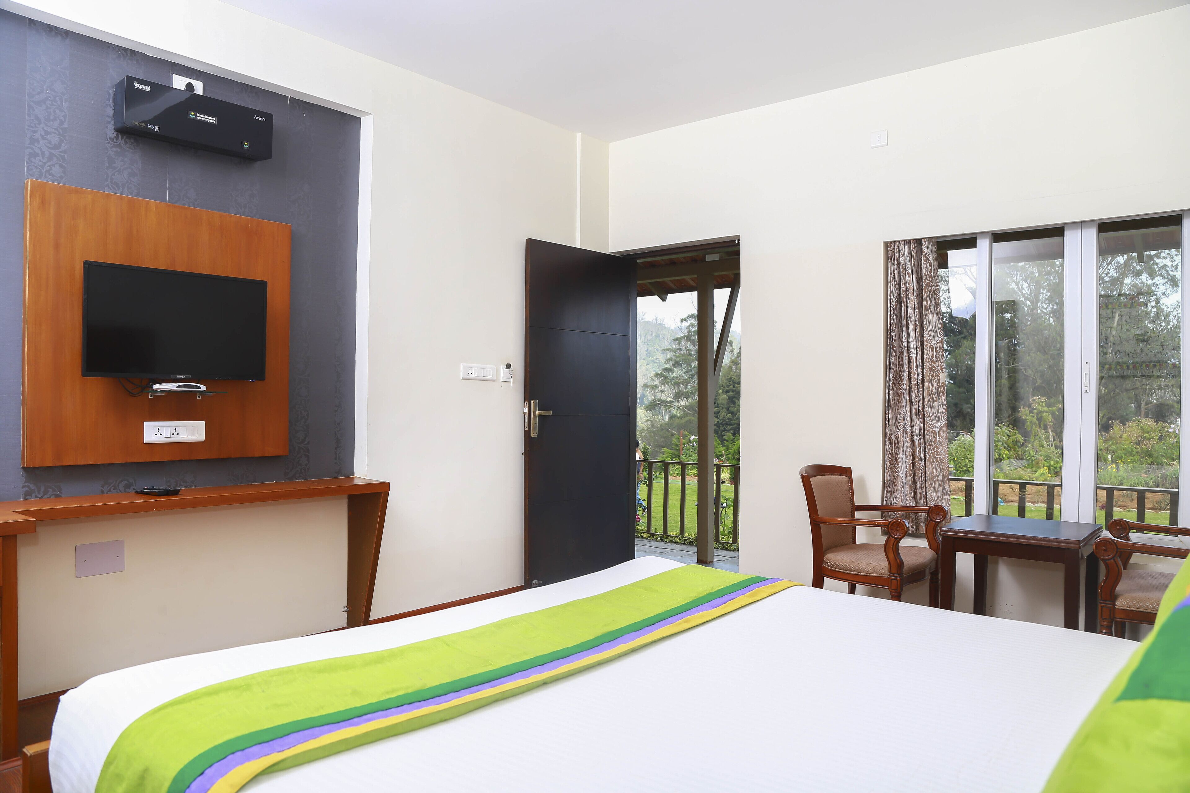 A hotel room with a bed, a TV mounted on the wall, a desk with a chair, and a balcony with a view of greenery.
