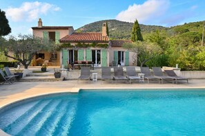 A pool with lounge chairs, a building with a tiled roof, and a mountainous backdrop.