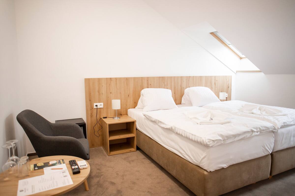 Ein Hotelzimmer mit einem Bett, einem Schreibtisch mit Lampe, einem Sessel und einem kleinen Tisch mit Fernbedienung und Unterlagen.