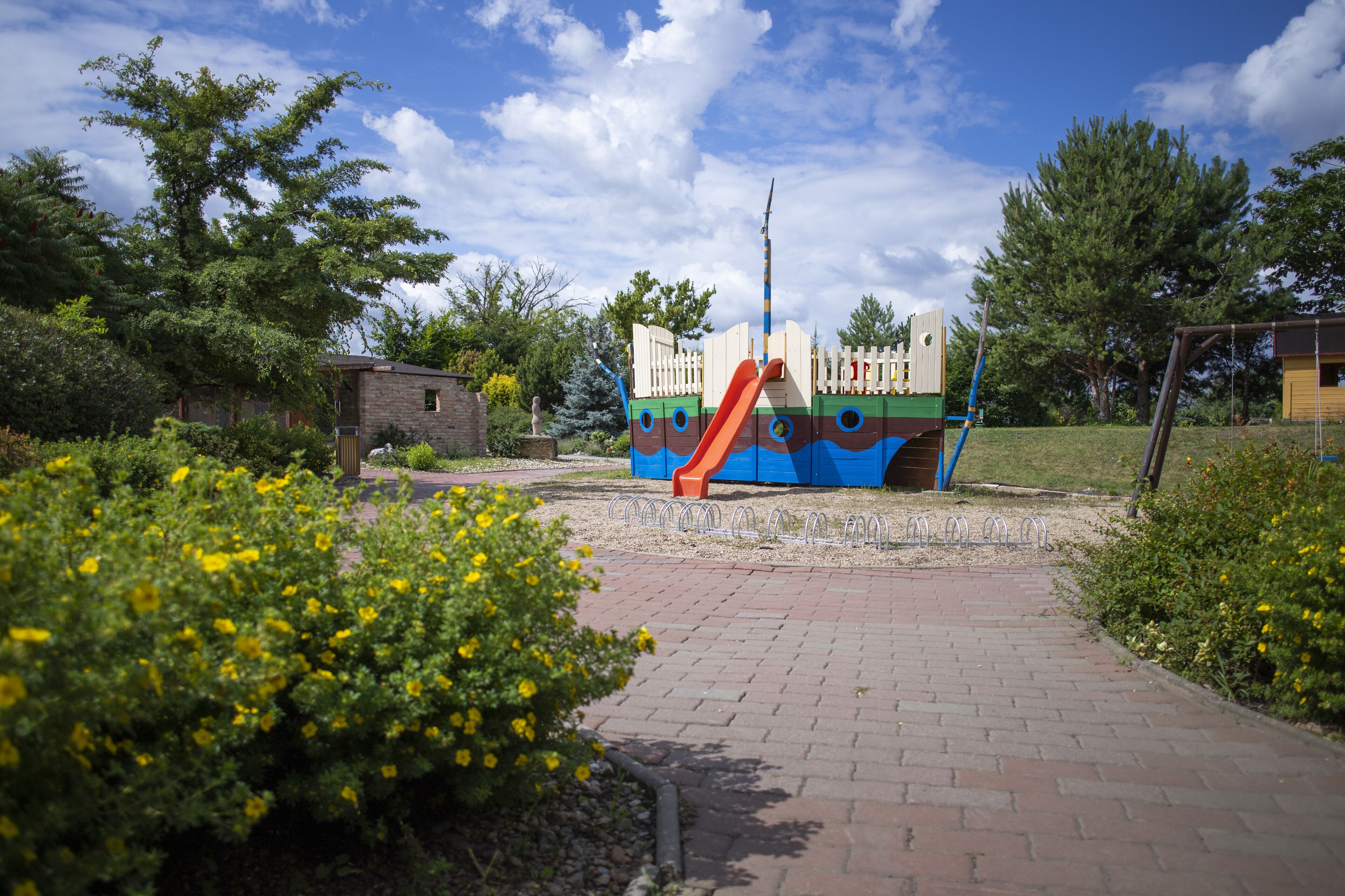 A playground with a slide and climbing structures, surrounded by greenery and a brick pathway.