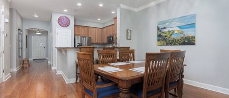 Un comedor con mesa y sillas de madera, una cocina al fondo y un cuadro en la pared.