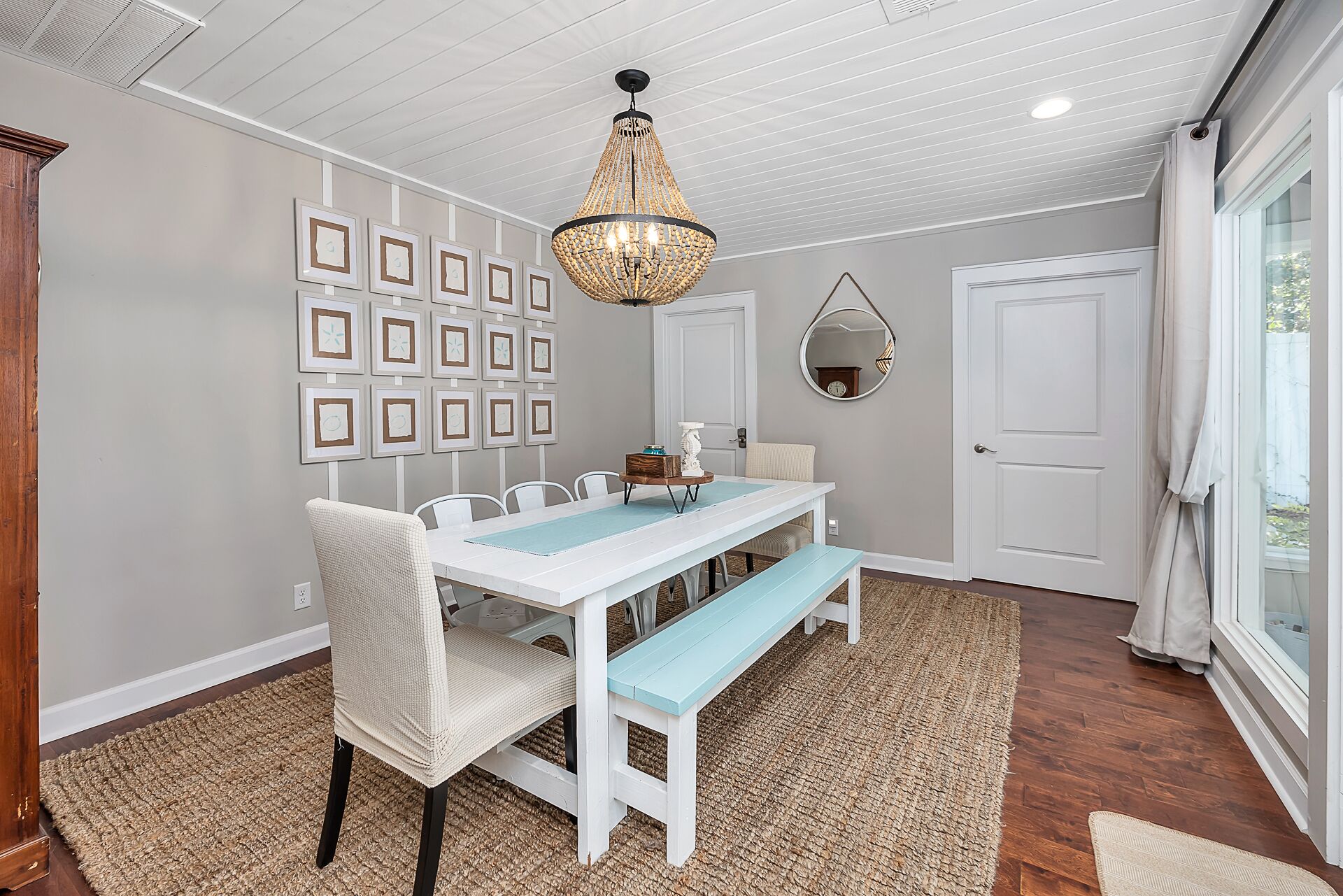 A dining area with a white table, blue bench, and a chandelier. There are framed pictures on the wall and a round mirror above the door.