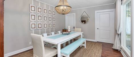 A dining area with a white table, blue bench, and a chandelier. There are framed pictures on the wall and a round mirror above the door.