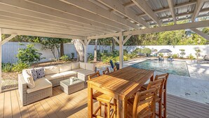 Una terrazza esterna con un tavolo e sedie in legno, un’area salotto imbottita e una piscina sullo sfondo.
