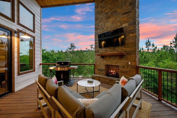 A covered deck with outdoor furniture, a stone fireplace, and a grill.
