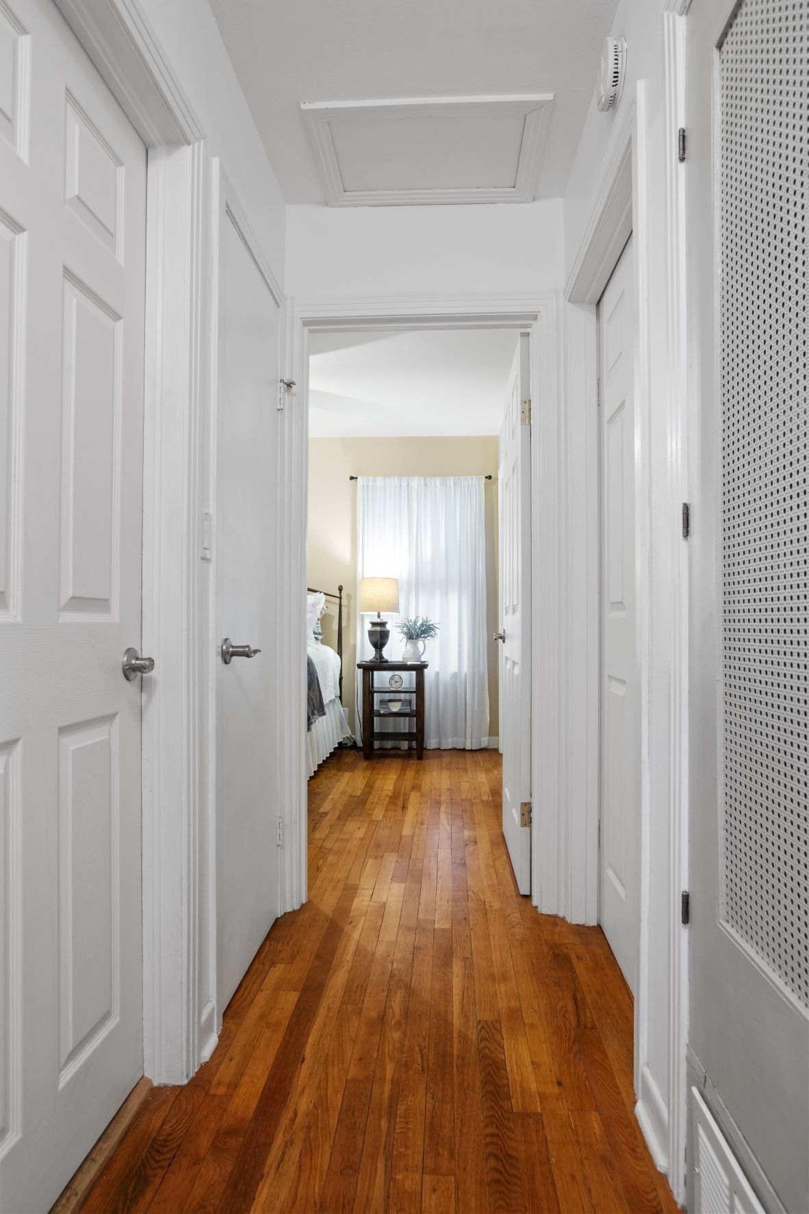 A hallway with white doors, wooden flooring, and a bed with a lamp in the background.